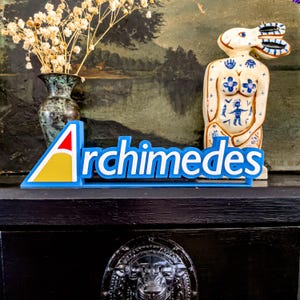 Puede incluir: Un letrero azul con la palabra "Archimedes" en blanco. El letrero tiene un logotipo rojo, amarillo y azul. Un jarrón decorativo con flores secas y una figura de cerámica están en el fondo.