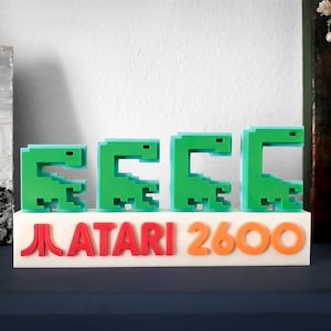 Puede incluir: Una pieza de exhibición retro de Atari 2600. El diseño presenta cuatro figuras verdes pixeladas con forma de dinosaurio sobre el logotipo de Atari y el texto "2600" en naranja. La base es blanca y la estética general recuerda a los videojuegos clásicos.