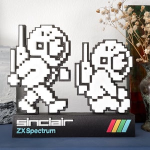 May include: A black and white 3D printed figurine of two characters from the video game Jet Set Willy, standing on a black base with the text "sinclair ZX Spectrum" and a rainbow stripe design.