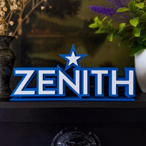 Pode incluir: Uma placa azul e branca impressa em 3D que diz "ZENITH". A placa apresenta uma base azul com letras brancas e uma estrela branca dentro de uma forma de estrela azul. É exibida em uma superfície preta.