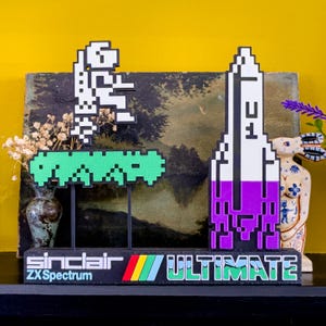 Peut inclure: Objet décoratif rétro avec des personnages pixélisés et le texte "Sinclair ZX Spectrum ULTIMATE". Le design comprend un personnage blanc, une plateforme verte et une fusée violette et blanche. La base est noire avec des rayures colorées.