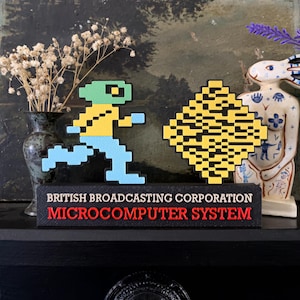 Peut inclure: Un objet décoratif de style rétro avec des personnages pixélisés et du texte. Les personnages incluent une personne en train de courir et une forme de diamant. Le texte indique "BRITISH BROADCASTING CORPORATION MICROCOMPUTER SYSTEM" en rouge et noir.