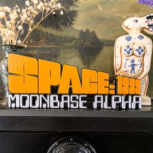 May include: A decorative sign with the words "SPACE 1999 MOONBASE ALPHA" in orange and silver lettering. The sign is displayed on a black surface, with a ceramic figurine of a rabbit in the background.