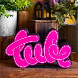 Könnte beinhalten: Ein dreidimensionales Schild mit dem Wort "Tude" in leuchtend pinker Schrift mit weißer Umrandung. Die Buchstaben sind in einer kursiven, stilisierten Schriftart verbunden und das gesamte Stück ist auf einem schwarzen Sockel montiert.