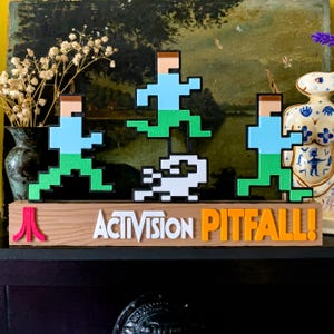 Puede incluir: Un letrero de madera con el texto "ACTIVISION PITFALL!" en blanco y naranja. Encima del letrero hay figuras pixeladas de un hombre corriendo en verde y azul, y una criatura blanca. Un logo rojo de Atari está a la izquierda.