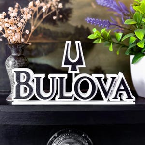 Op de afbeelding: Een decoratief Bulova logo-bord in wit en zwart. Het bord toont de merknaam in een vetgedrukte letter en een opvallend symbool. Het staat op een zwart oppervlak, met planten op de achtergrond, wat een stijlvolle presentatie creëert.