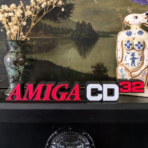 May include: A decorative sign with the words "AMIGA CD32" in red and white block letters. The sign is displayed on a black surface, with a vintage vase and a ceramic figurine in the background.
