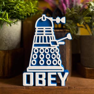 Könnte beinhalten: Ein blau-weißes 3D-gedrucktes Schild mit einer stilisierten Dalek-Figur über dem Wort OBEY in fetten Blockbuchstaben. Das Design verwendet eine geschichtete Ästhetik mit einer weißen Umrandung und einer blauen Basis für einen Kontrast.