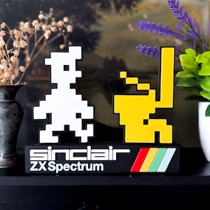Peut inclure: Objet décoratif rétro avec des personnages pixélisés blancs et jaunes. La base noire porte l'inscription "Sinclair ZX Spectrum" et un motif rayé coloré. Idéal pour les fans de jeux vidéo rétro.