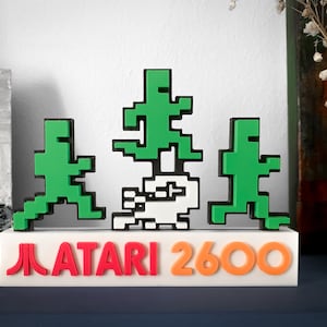 Puede incluir: Una base blanca con el texto "ATARI 2600" en letras rojas y naranjas. Tres figuras pixeladas verdes se encuentran sobre la base, con una figura pixelada blanca en el centro.
