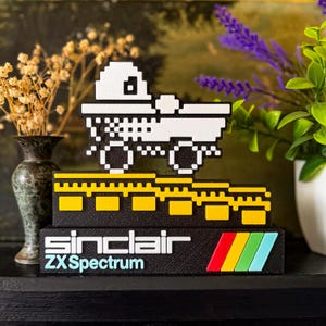 Könnte beinhalten: Ein pixeliger LKW in Schwarz und Weiß steht auf einem schwarzen Sockel mit gelben Akzenten. Der Sockel zeigt "sinclair ZX Spectrum" mit einem farbenfrohen Streifen. Ein Sammlerstück für Retro-Gaming.