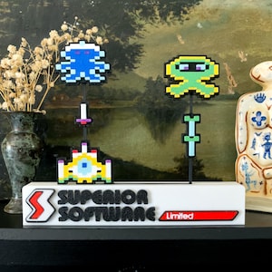 May include: A white base with the text "SUPERIOR SOFTWARE Limited" supports pixelated video game characters. The characters include a blue octopus, a green alien, and a yellow and white spaceship. The background features a vase with dried flowers and a decorative statue.