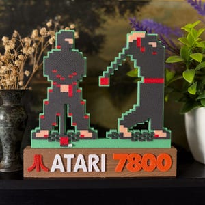 May include: A pixelated Atari 7800 game scene featuring two black ninja figures with red accents, standing on a green base. The Atari 7800 logo is displayed on a wooden block below the figures. The figures are approximately 5 inches tall.