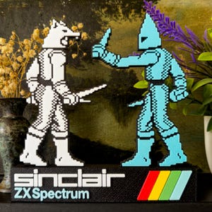 Op de afbeelding: Een retro sculptuur met twee gepixelde figuren in gevecht. Eén figuur is wit, de andere lichtblauw. De zwarte basis toont "Sinclair ZX Spectrum" en een kleurrijk streepontwerp. Een nostalgisch item voor gamers.