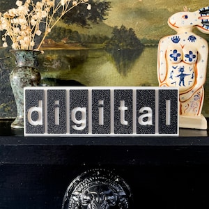 Può includere: Un cartello in bianco e nero che dice la parola "digital". Il cartello è composto da singoli blocchi rettangolari neri con lettere bianche. Il cartello è su una superficie nera, con un vaso di fiori secchi e una statuina decorativa sullo sfondo.
