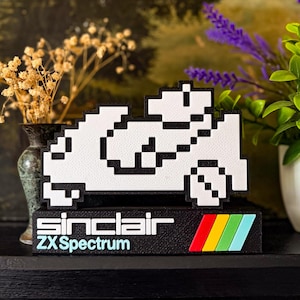 May include: A white and black pixelated car sculpture with the text "Sinclair ZX Spectrum" on a black base. The base features a colourful stripe design. The sculpture is displayed on a dark surface.