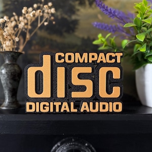 Op de afbeelding: Een driedimensionaal bord met de tekst COMPACT DISC DIGITAL AUDIO. Het woord DISC is weergegeven in grote, getextureerde gouden letters tegen een zwarte achtergrond, met de woorden COMPACT en DIGITAL AUDIO in kleinere gouden letters.