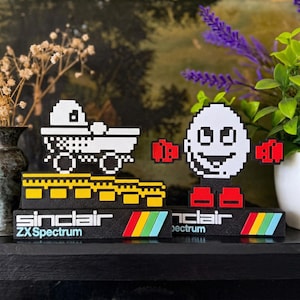 Könnte beinhalten: Pixelige Kunstwerke mit einem weißen LKW und einem lächelnden, eiförmigen Charakter. Beide stehen auf schwarzen Sockeln mit dem Text "Sinclair ZX Spectrum" und einem Regenbogenstreifen. Inspiriert von Retro-Videospielen.