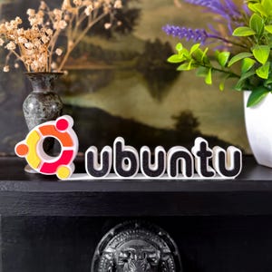 Op de afbeelding: Een kleurrijk Ubuntu-logo en woordmerk. Het woord 'Ubuntu' is in witte letters met zwarte vulling. Het logo heeft oranje, gele en rode accenten. Het item wordt weergegeven op een zwart oppervlak, met planten en een vaas op de achtergrond.