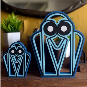 Peut inclure: Deux objets d'art décoratifs bleus et noirs avec un motif de hibou stylisé. La pièce la plus grande mesure environ 15 cm de haut, et la plus petite environ 8 cm. Les figures de hibou ont des yeux blancs et sont posées sur une surface en bois.