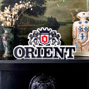Pode incluir: Uma placa preta e branca "ORIENT" com um brasão com dois leões, uma coroa e um escudo vermelho. A placa é exibida em uma superfície preta, com um vaso de flores secas e uma estatueta de cerâmica no fundo.