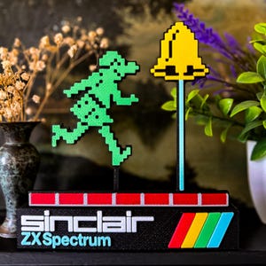 Peut inclure: Une pièce d'exposition rétro Sinclair ZX Spectrum. Elle présente une figurine verte pixélisée courant vers une cloche jaune. La base noire porte le logo Sinclair et le texte ZX Spectrum, ainsi qu'un design à rayures rouges, blanches et colorées.