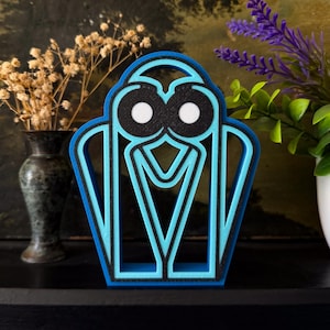Peut inclure: Un objet décoratif bleu et noir avec un design stylisé. L'objet présente un contour bleu clair et un centre noir. Le design comprend deux cercles blancs et un élément en forme de V.