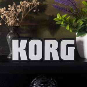 Peut inclure: Panneau KORG noir et blanc. Les lettres blanches ont une surface texturée, soulignées de noir. Le panneau est affiché sur une surface noire, avec des vases décoratifs et des plantes en arrière-plan.