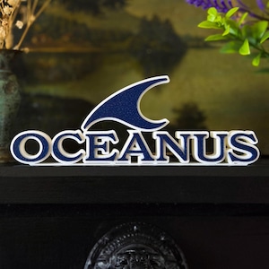 May include: A white and navy blue "OCEANUS" sign with a stylized wave design. The letters are outlined in navy blue with a white border. The wave design is navy blue with a white outline. The sign is displayed on a black surface.