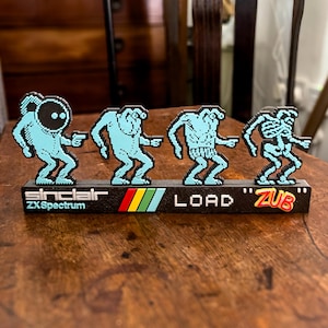 Può includere: Una statuetta 3D stampata in blu e nero che rappresenta un computer Sinclair ZX Spectrum che carica un gioco. La statuetta presenta quattro personaggi pixelati e il testo "LOAD "ZUB"".