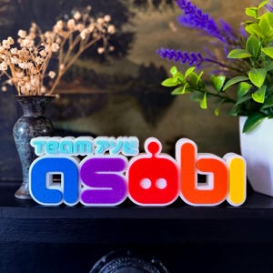 May include: A colorful, 3D-printed sign that spells out "asobi" in vibrant hues of blue, purple, red, orange, and yellow. The word "Team" is above the word "asobi" in a teal color. The sign is displayed on a black surface.