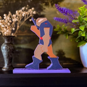 May include: A wooden figurine of a character in a walking pose. The figure has a blue and orange color scheme with tan arms and a purple base. The figurine is standing on a purple rectangular base.