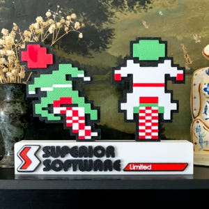 May include: A white base with the words "SUPERIOR SOFTWARE Limited" in black and red. Two pixelated figures in green, red, and white stand on top. One figure is riding a pixelated vehicle. The other figure is falling.