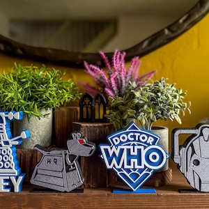 May include: Four 3D-printed decorative figures featuring a blue and white Dalek with the text OBEY, a grey K-9 robot dog, a blue and white Doctor Who logo sign, and a grey Cyberman head. These items are arranged on a wooden surface.