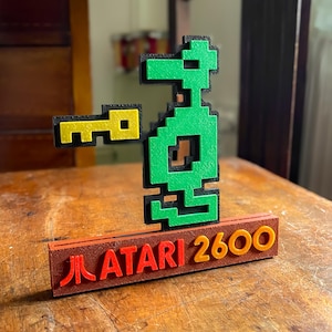 Może przedstawiać: Zielona postać z pikseli trzymająca żółty klucz, stojąca na brązowej podstawie z napisem "ATARI 2600" w kolorze czerwonym i pomarańczowym.