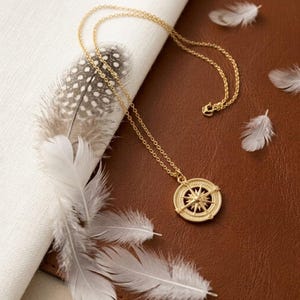 May include: A gold-colored compass pendant necklace with a delicate chain. The pendant features a detailed compass rose design. The necklace is displayed with white feathers and a patterned feather on a brown surface.