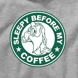 Puede incluir: Una sudadera gris con un gráfico circular verde que presenta un personaje de dibujos animados somnoliento y el texto "SLEEPY BEFORE MY COFFEE".