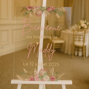 Op de afbeelding: Een helder acrylen welkomstbord op een witte ezel, versierd met bloemstukken. De roze tekst luidt "Bienvenue au baptême de Maddy Le 12 juillet 2025". De bloemstukken bevatten roze rozen en gedroogde grassen.