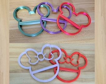 May include: Four plastic cookie cutters in the shape of Mickey Mouse and Minnie Mouse. Two are green, one is purple, one is red, one is white, and one is red.