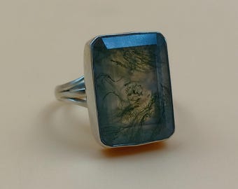 May include: A rectangular moss agate gemstone ring with a silver band. The gemstone has a translucent appearance with green and brown inclusions. The ring is set on a neutral background.