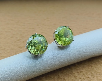 May include: A pair of round, faceted, light green gemstone stud earrings. The gemstones are set in a silver-colored metal. The earrings are displayed on a white surface against a neutral background.