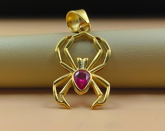 May include: Gold-colored spider pendant with a teardrop-shaped, red gemstone at its center. The pendant has a bail for a chain and is set against a neutral background. The spider design features intricate, geometric legs.
