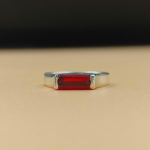 May include: A silver ring with a rectangular, deep red gemstone. The gemstone is set horizontally in the center of the ring. The ring is displayed on a light brown surface, with a dark background.