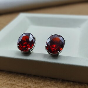 May include: A pair of oval-cut, deep red gemstone stud earrings. The gemstones are set in a silver-colored metal with four prongs. The earrings are displayed on a white surface.