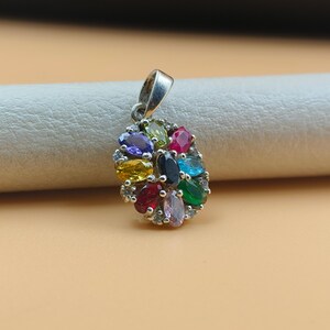 May include: Silver pendant with an oval cluster of colorful gemstones. The pendant features a variety of jewel tones, including purple, yellow, red, green, and blue. The gemstones are set in a silver setting.