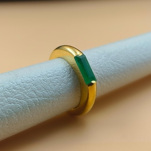 Puede incluir: Un anillo de oro con una piedra preciosa rectangular de color verde esmeralda. El anillo tiene un diseño simple y moderno con un acabado pulido. La piedra preciosa está engastada horizontalmente en el centro de la banda.