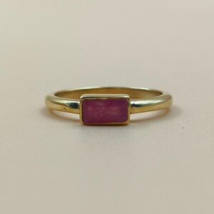 May include: A gold ring featuring a rectangular, pink gemstone. The ring has a smooth, rounded band, and the gemstone is set in a rectangular bezel. The ring is shown on a light pink surface.