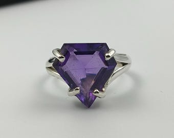 May include: A silver ring featuring a large, faceted, deep purple gemstone. The gemstone is cut into a unique geometric shape. The ring has a split shank design, with the band splitting into two separate strands on each side of the gemstone.
