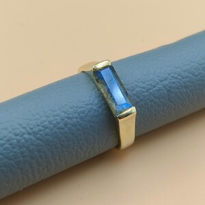 May include: A gold ring with a rectangular, blue gemstone. The ring is set on a gray surface, with the gold band and gemstone catching the light. The gemstone has a faceted appearance.