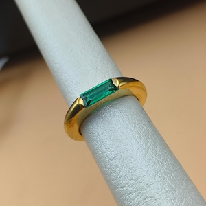 Könnte beinhalten: Ein goldener Ring mit einem rechteckigen, smaragdgrünen Edelstein. Der Ring wird auf einer weißen Oberfläche präsentiert, die das polierte Goldband und das leuchtende Grün des Steins hervorhebt. Das Design des Rings ist schlicht und elegant.